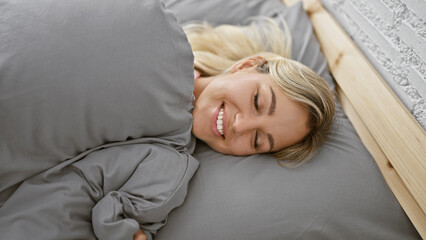 Obraz premium Smiling young adult caucasian woman relaxing in a cozy bedroom with grey bedding, portraying comfort and happiness.