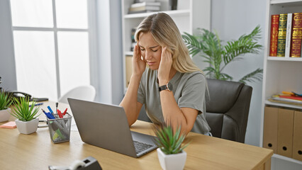 A stressed young woman experiencing headache while working on her laptop in a modern office setting