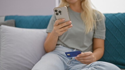 A young woman sits indoors, holding a credit card and smartphone while shopping online from home.