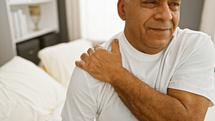 A middle-aged hispanic man in a white shirt expressing discomfort or pain while sitting on a bed in...