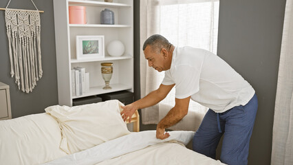 A middle-aged hispanic man makes the bed in a well-organized bedroom interior, reflecting a sense...