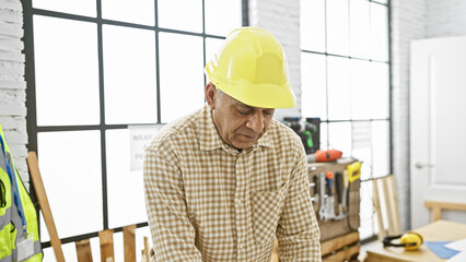 Mature hispanic man in a workshop wearing a hardhat focused on carpentry work