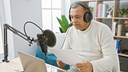 Hispanic mature man with headphones narrating script in a modern radio studio setup indoors.