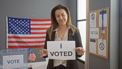 Smiling woman holding 'i voted' sign in a polling station with american flag.