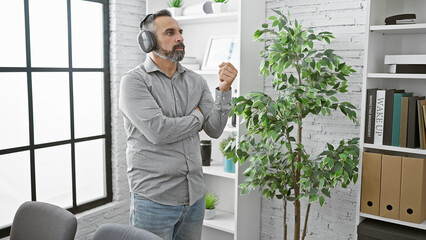 A bearded senior hispanic man enjoying music with headphones in a modern office, exuding casual professionalism.