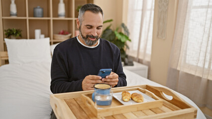 Mature man with beard using smartphone in bedroom with breakfast tray.