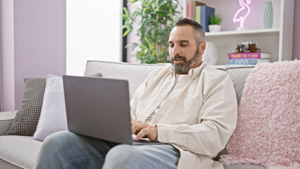 A mature hispanic man with a beard and grey hair using a laptop on a sofa in a cozy living room.