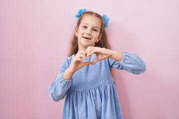 Young little girl standing over pink background smiling in love showing heart symbol and shape with hands. romantic concept.