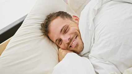 A handsome adult man with a beard smiling while lying comfortably in white bed sheets in a home bedroom setting.
