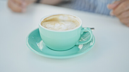 Close-up of a frothy cappuccino in a teal cup with a blurred adult man in the background at an outdoor cafÃ©.