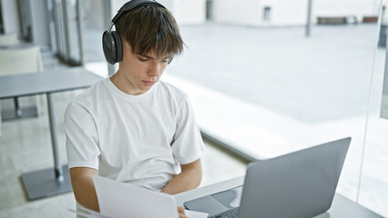 A young caucasian male teenager studies at a library table with a laptop and headphones.