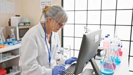 A middle-aged woman scientist in a lab coat working on a computer in a bright laboratory setting.