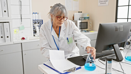Mature woman scientist in lab analyzes data on computer with flask, beaker and clipboard in a...