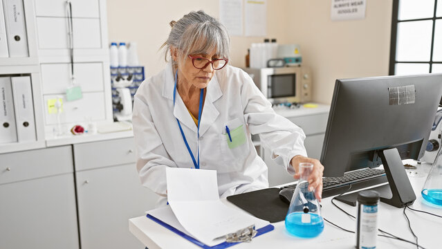 Mature woman scientist in lab analyzes data on computer with flask, beaker and clipboard in a clinical research environment. - Powered by Adobe