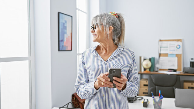 Mature woman with grey hair holding a smartphone in a modern office, embodying professionalism and experience in a workplace setting.