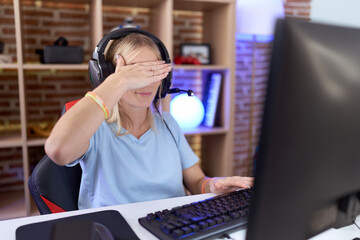 Young caucasian woman playing video games wearing headphones covering eyes with hand, looking...