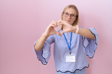 Young caucasian business woman wearing id card smiling in love doing heart symbol shape with hands....