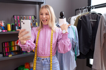 Young caucasian woman dressmaker designer on video call with smartphone pointing thumb up to the side smiling happy with open mouth