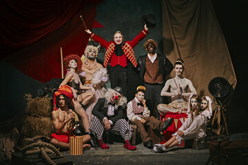 Diverse cast of vintage circus performers poses over dark retro circus backstage background....