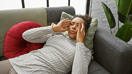 A stressed young woman with curly hair lying on a couch indoors touching her forehead in a home...