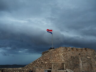 雨雲が迫るフヴァル城塞にはためく国旗 - obrazy, fototapety, plakaty