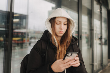 Half length portrait of girl in casual wear holding smartphone for blogging in the city in the autumn-winter season. Teen surfing the internet outdoors.