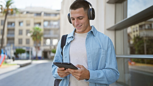 Smiling young hispanic man in casual wear using a tablet on a sunny urban street.