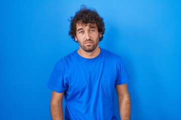 Hispanic young man standing over blue background looking sleepy and tired, exhausted for fatigue and hangover, lazy eyes in the morning. - obrazy, fototapety, plakaty