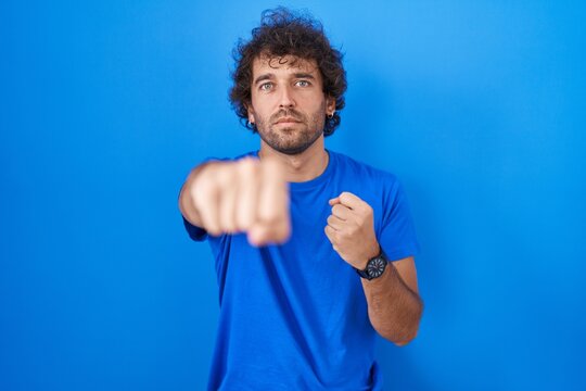 Hispanic young man standing over blue background punching fist to fight, aggressive and angry attack, threat and violence