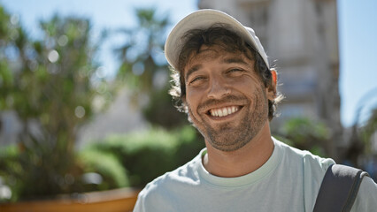 Handsome smiling hispanic man with beard and green eyes wearing a cap outdoors in urban setting