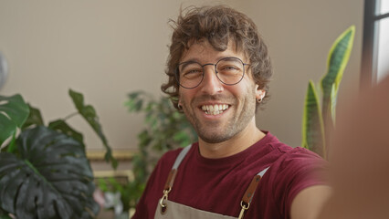 A cheerful man with glasses and a beard takes a selfie in a green plant-filled flower shop, exuding warmth and friendliness.