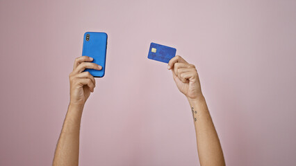 Man's tattooed arms holding smartphone and credit card against pink wall, depicting online payment or shopping.