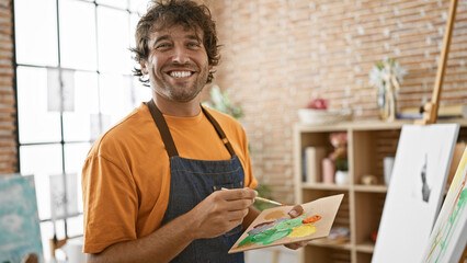 A smiling hispanic man painting on a canvas in a bright, artistic studio setting.