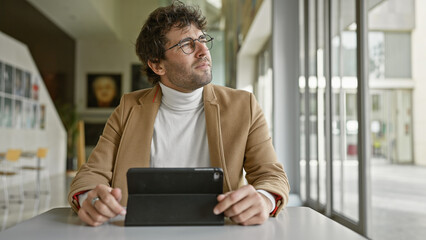Thoughtful hispanic man with glasses in a modern office holding tablet