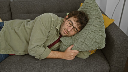 A young man with glasses sleeps cozily with a green pillow on a gray couch at home.