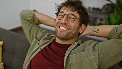Handsome hispanic man with glasses and beard smiling while relaxing on a couch indoors.