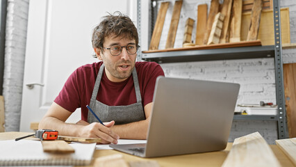 Bearded craftsman in apron working on laptop in a woodshop, surrounded by timber and tools.