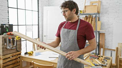 Hispanic man inspects wood in a sunny carpentry workshop wearing apron and glasses.