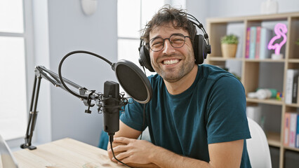 Hispanic man with headphones smiling in radio studio setup with microphone and pop shield