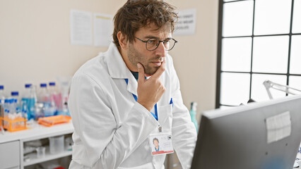 Thoughtful hispanic man with beard working at computer in hospital laboratory, portraying healthcare professional.