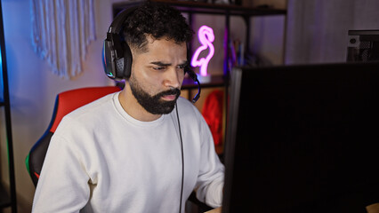 Focused hispanic man with beard wearing headphones in a neon-lit gaming room at night.