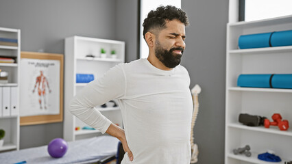 A pained young hispanic man with a beard grimaces while holding his lower back inside a modern physiotherapy clinic. - obrazy, fototapety, plakaty