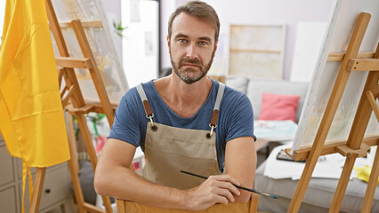 Portrait of a confident middle-aged man, artist, indoors, holding a paintbrush and wearing an apron.
