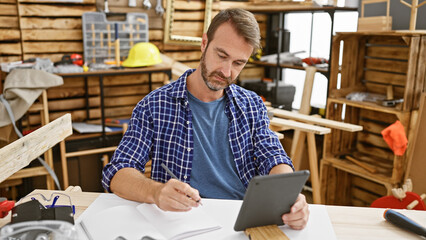 Bearded craftsman in blue shirt using tablet and writing in a bright woodworking workshop