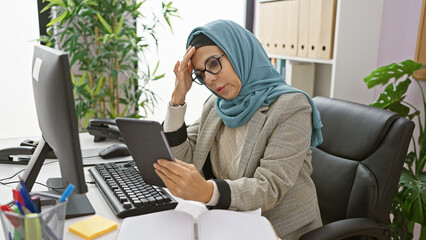 A mature, hijab-wearing woman looks seriously at a tablet in a modern office, exuding professionalism and focus.