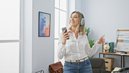 A joyful middle-aged woman enjoys music on headphones while holding a smartphone in a modern office.