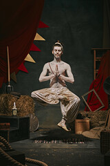 Pale man, yogi performing over dark retro circus backstage background. Magic and illusion festival....
