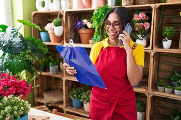 Young beautiful arab woman florist talking on smartphone reading clipboard at flower shop