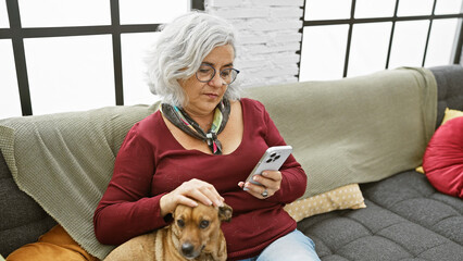 Mature woman relaxes at home, petting her dog while browsing her smartphone on a comfortable sofa...