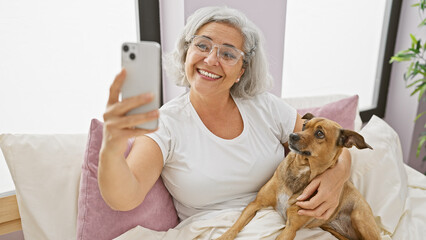 A grey-haired woman takes a selfie with her dog in a bright bedroom setting